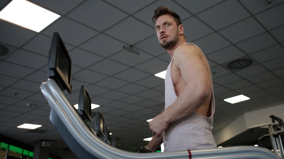 Low angle of muscular adult male athlete in sportswear during training on running machine in modern spacious fitness center while looking away